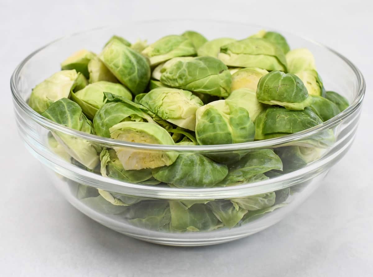 An image of Brussels sprouts that are sliced in halves and quarters in a glass bowl set on a white table.