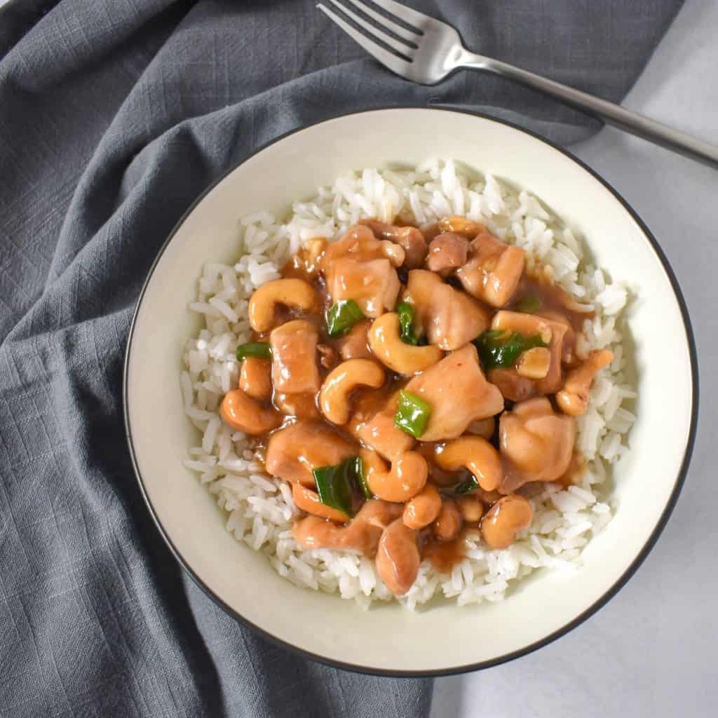 An image of the cashew chicken served over white rice in a white bowl with a black rim. The bowl is set on a white table with a gray linen and a fork to the top right side.