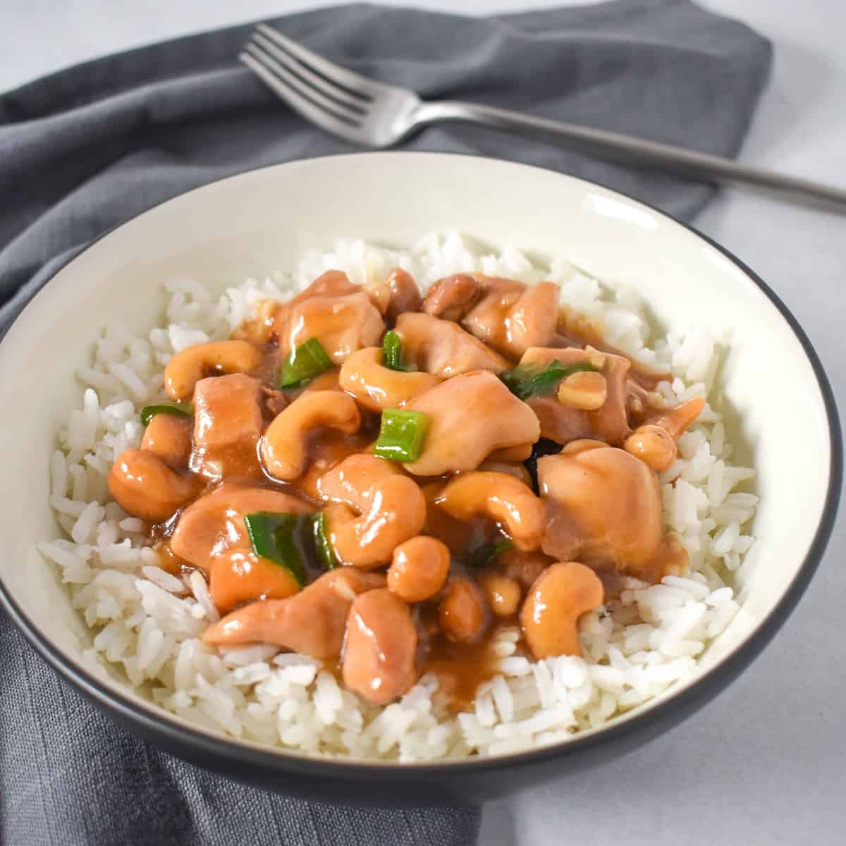 The chicken cashew served on white rice in a white bowl with a black rim. The bowl is set on a white table with a gray linen and a fork.