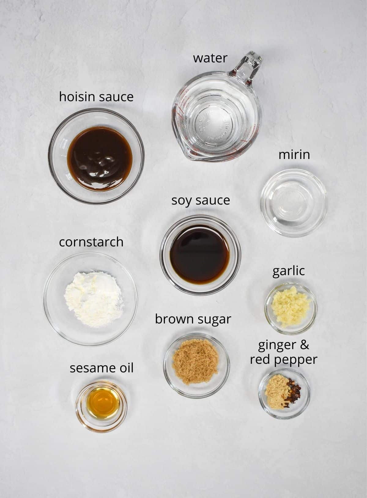 The ingredients for the sauce arranged in glass bowls and set on a white table. Each ingredient has a small label with the name in black letters.