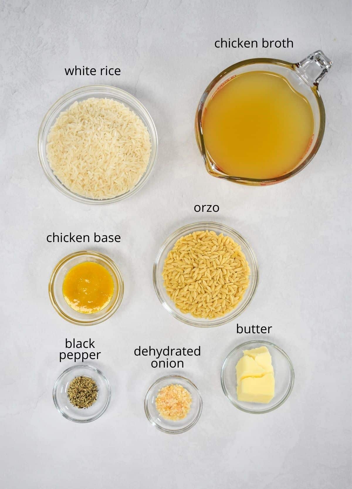 The ingredients for the rice arranged in glass bowls, set on a white table. Each ingredient has a small label with the name above it in small black letters.