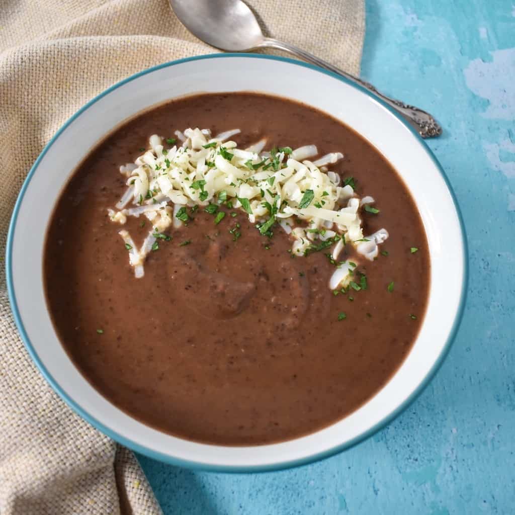 The creamy black bean soup served in a white bowl with a light blue rim and garnished with shredded Monterey jack cheese and chopped parsley. The bowl is set on a light blue table with a beige linen and a spoon to the top right side.