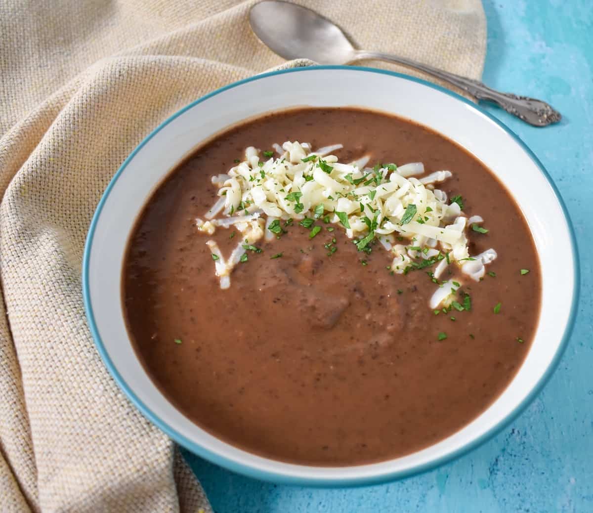 A close up image of the soup served in a white bowl with a light blue rim. The soup is garnished with shredded Monterey jack cheese and parsley. The bowl is set on a light blue table with a beige linen and a spoon to the top right side.