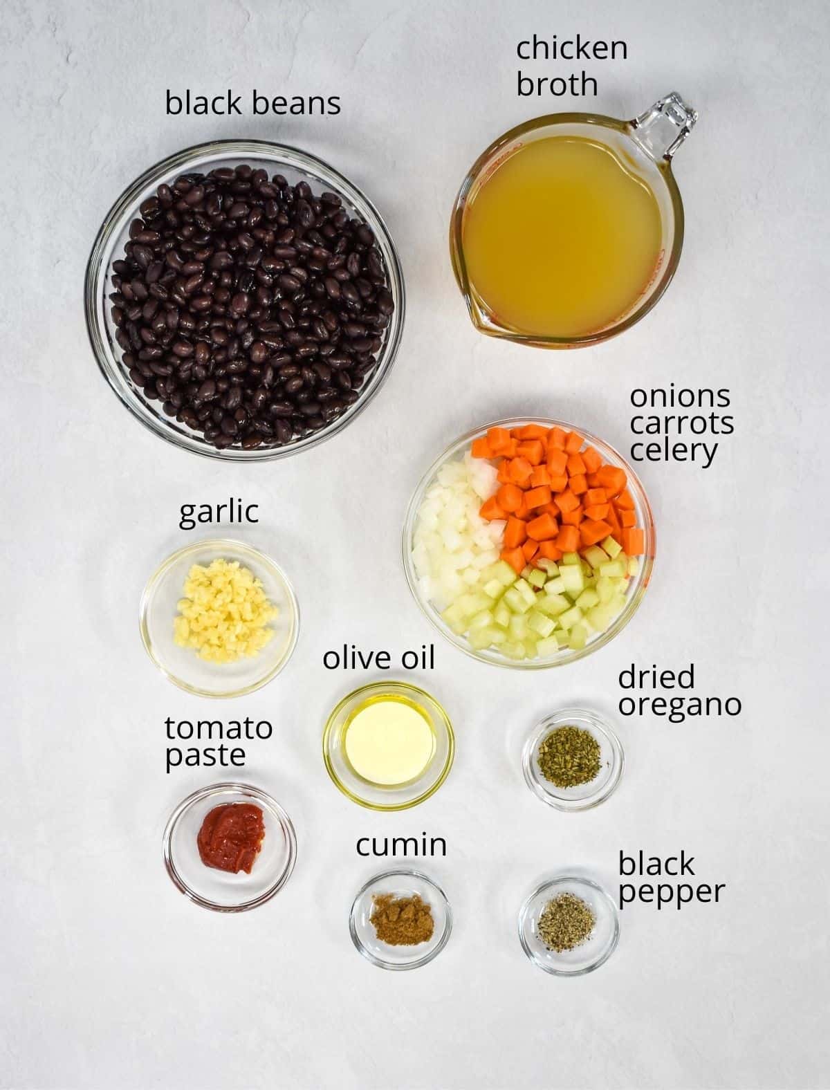 The ingredients for the black bean soup arranged in glass bowls and set on a white table. Each ingredient has a small label with the name above it in small black letters.
