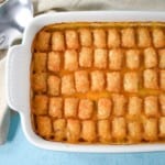 An image of the ground turkey tater tot casserole in a white casserole dish set on a light blue table with a beige linen and a serving spoon.