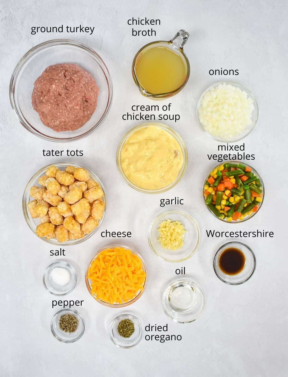The ingredients for the tater tot casserole prepped and arranged in glass bowls and set on a white table. Each ingredient has a label with the name above it in small black letters.
