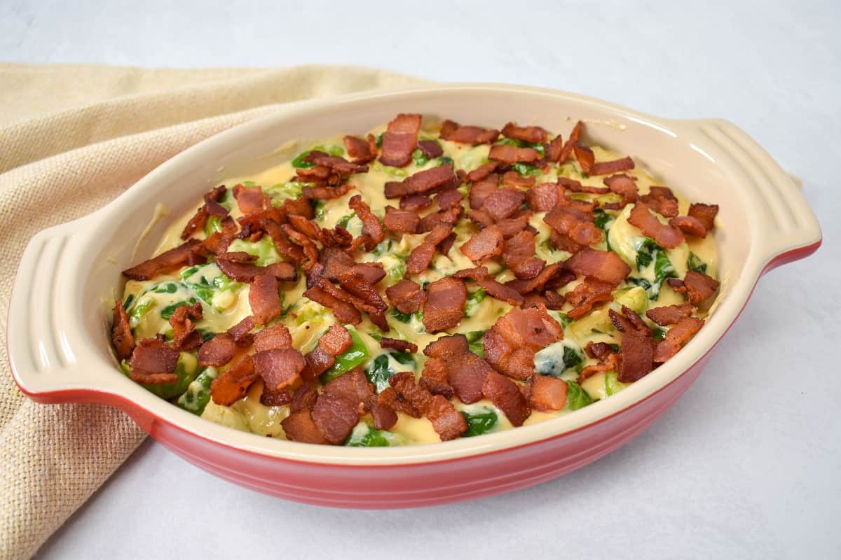 An image of the Brussels sprouts combined with the cheese sauce and topped with the crispy bacon in a casserole dish set up before baking. The dish is red with an off-white inside. 