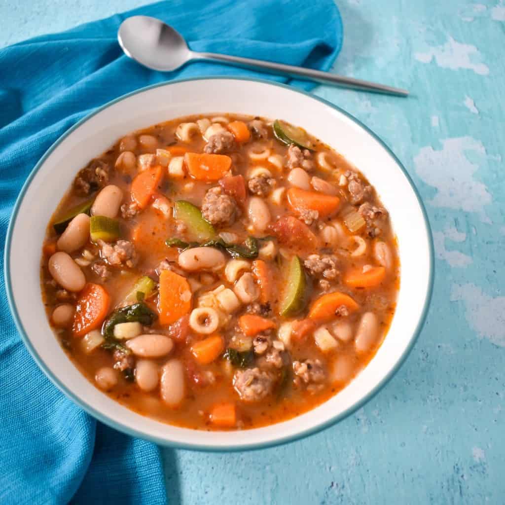 The sausage minestrone soup served in a white bowl with an aqua rim. The bowl is set on a light aqua table with an aqua linen and a spoon.