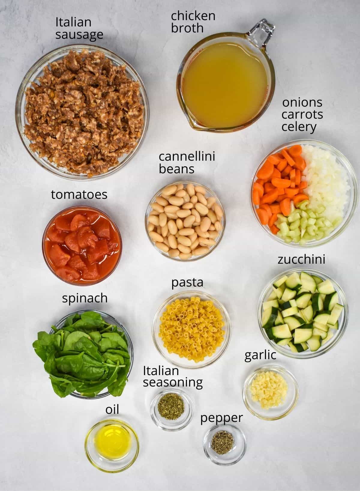 The ingredients for the soup prepped and arranged in glass bowls on a white table. Each ingredient has a small label with the name in black letters.