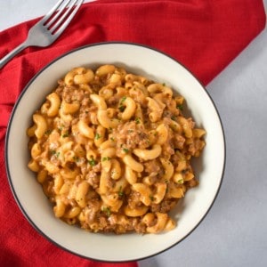 The cheeseburger macaroni served in a white bowl with a black rim. It is set on a white table with a red linen and fork.