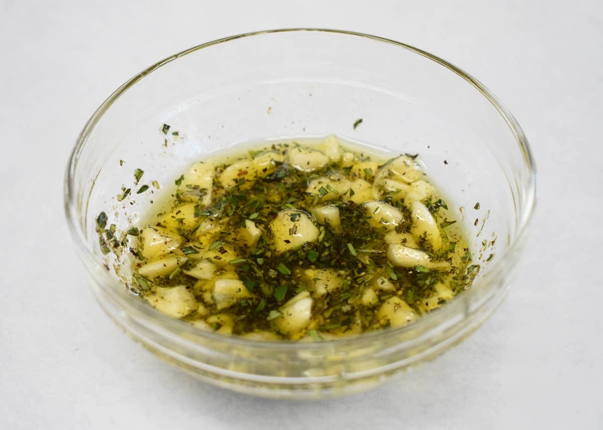 An image of the olive oil, garlic, herbs and spices mixed together in a glass bowl that is set on a white table.