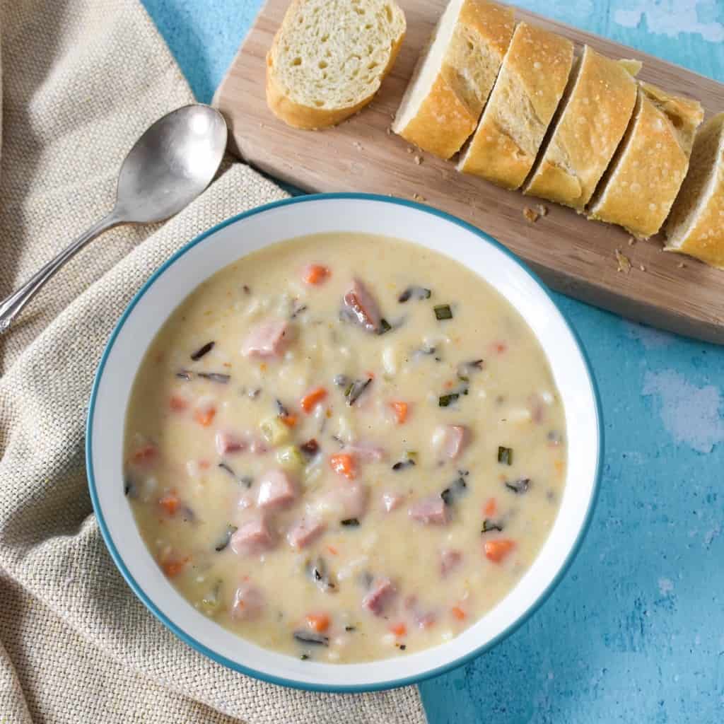 The ham and wild rice soup served in a white bowl with an aqua rim on a light blue table. To the top right side is a small wood cutting board with sliced bread loaf and to the right is a spoon and a beige linen.