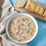 The ham and wild rice soup served in a white bowl with an aqua rim on a light blue table. To the top right side is a small wood cutting board with sliced bread loaf and to the right is a spoon and a beige linen.