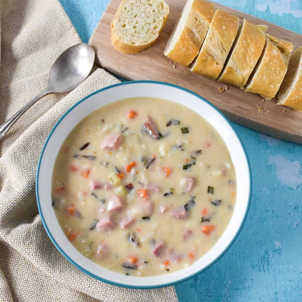 The ham and wild rice soup served in a white bowl with an aqua rim on a light blue table. To the top right side is a small wood cutting board with sliced bread loaf and to the right is a spoon and a beige linen.