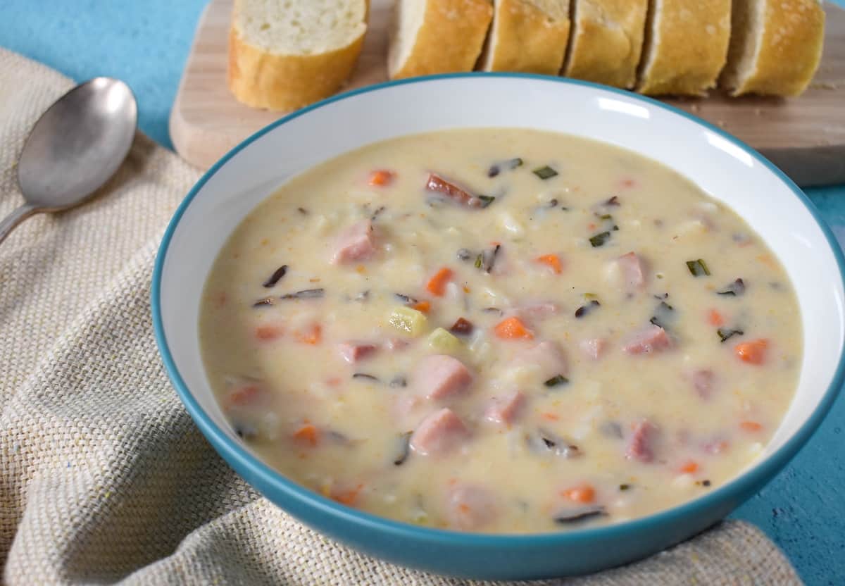 A close up image of the soup served in a white bowl with an aqua rim. There is a sliced bread loaf in the background and a spoon and beige linen to the left side.