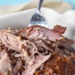 A close up image of the slow roasted pork shoulder in a white baking dish with a portion being held up by a large serving fork.