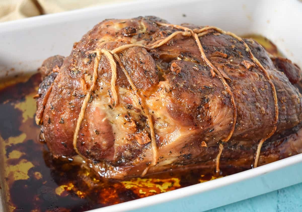 The cooked pork still tied with twine and in the drippings in a white baking dish.