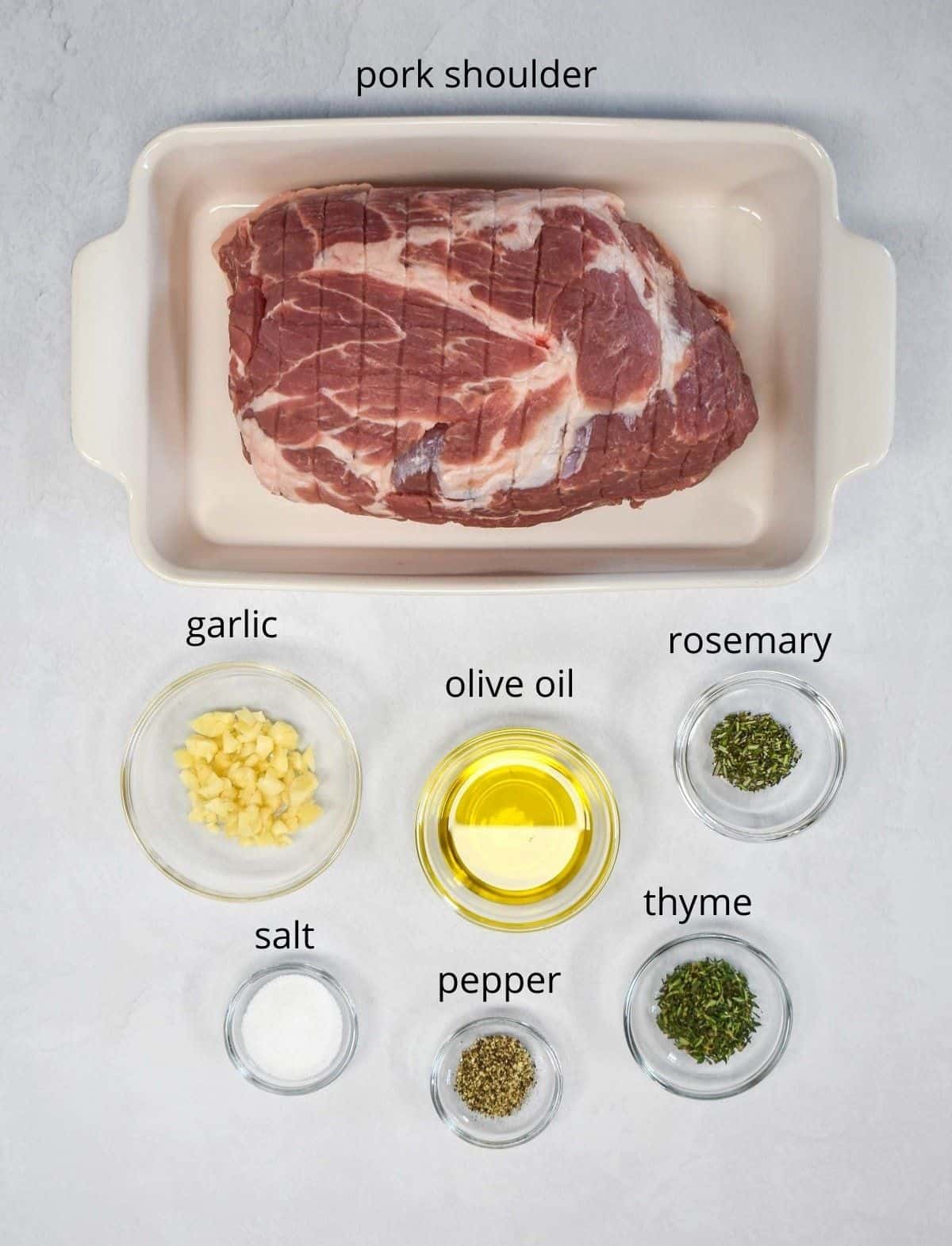 An image of the ingredients for the pork arranged on a white table. The pork is in a white baking dish and the rest of the ingredients are in glass bowls. Everything is set on a white table and each item has the name on top in small black letters.