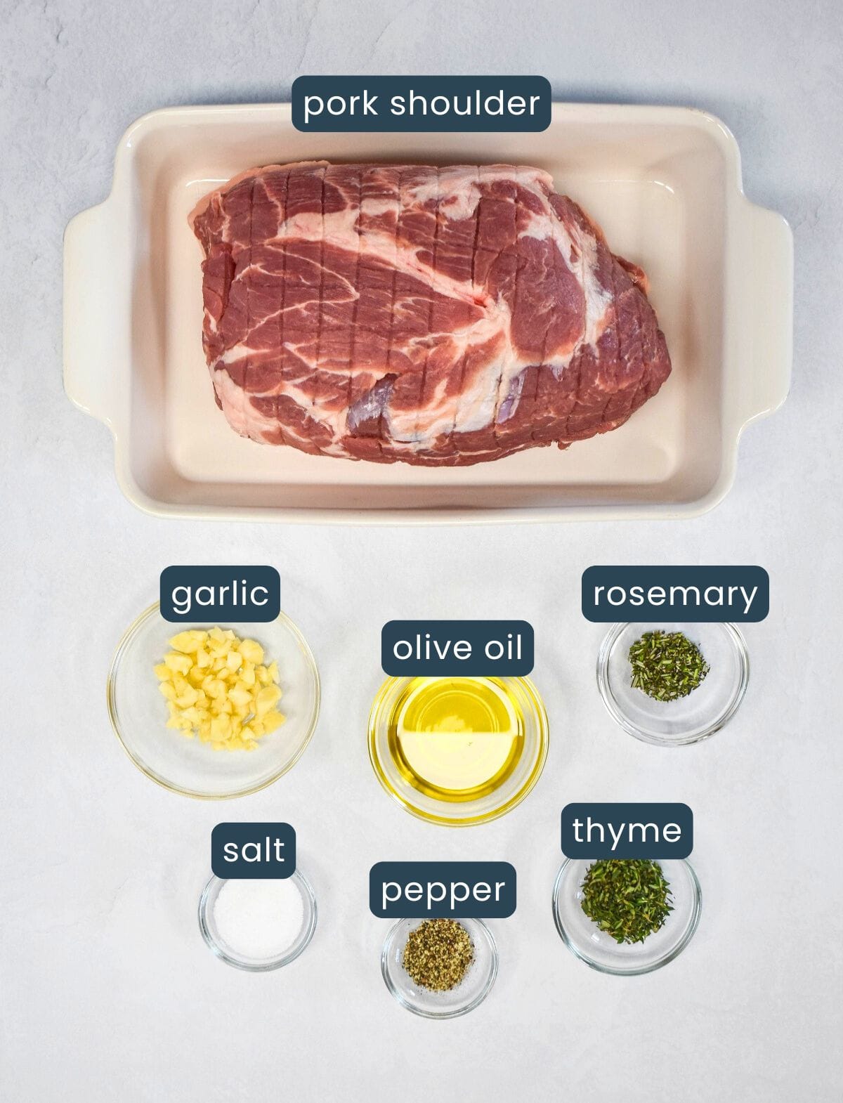 Ingredients for slow roasted pork shoulder displayed in small glass bowls on a light surface. Includes a large boneless pork shoulder in a white baking dish, and separate bowls with garlic, olive oil, rosemary, thyme, salt, and pepper.
