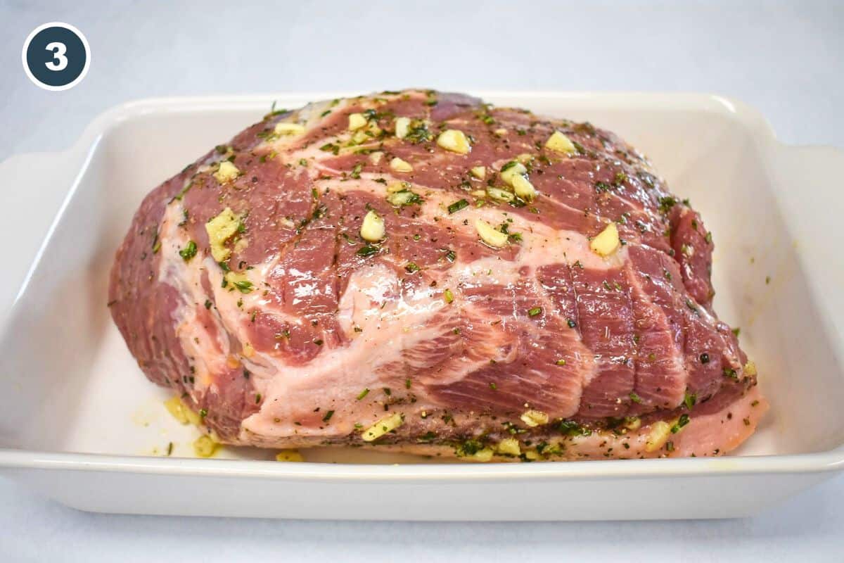 Pork shoulder coated with garlic herb mixture in a baking dish.