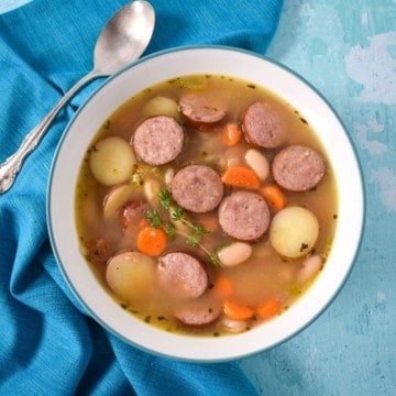 The smoked sausage soup served in a white bowl with an aqua rim. The bowl is set on a light blue table with an aqua linen and spoon to the left side.