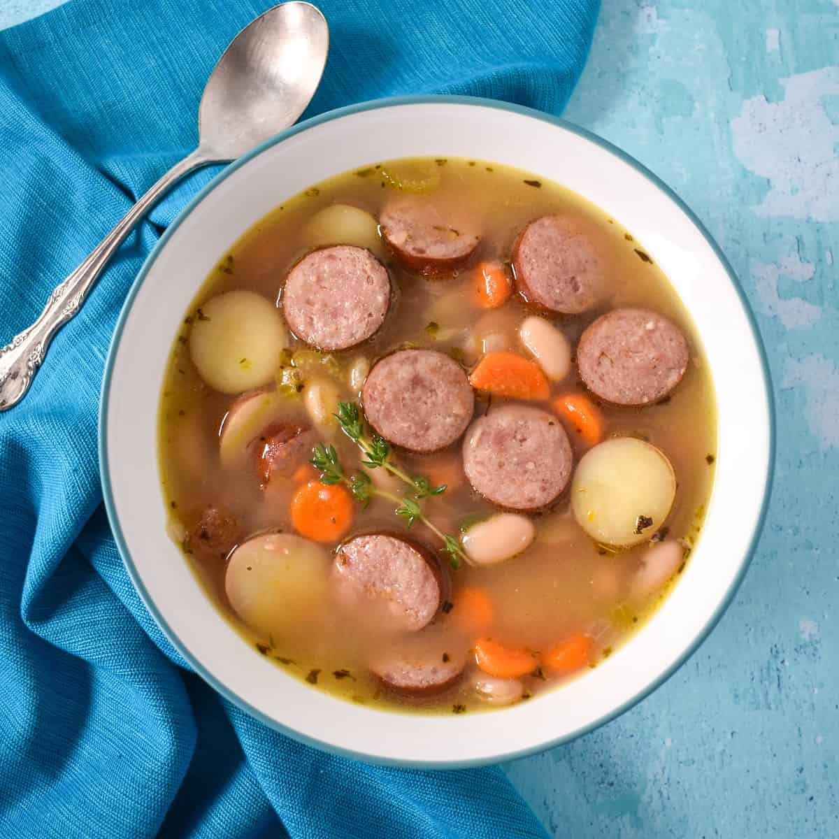 The smoked sausage soup served in a white bowl with an aqua rim. The bowl is set on a light blue table with an aqua linen and spoon to the left side.