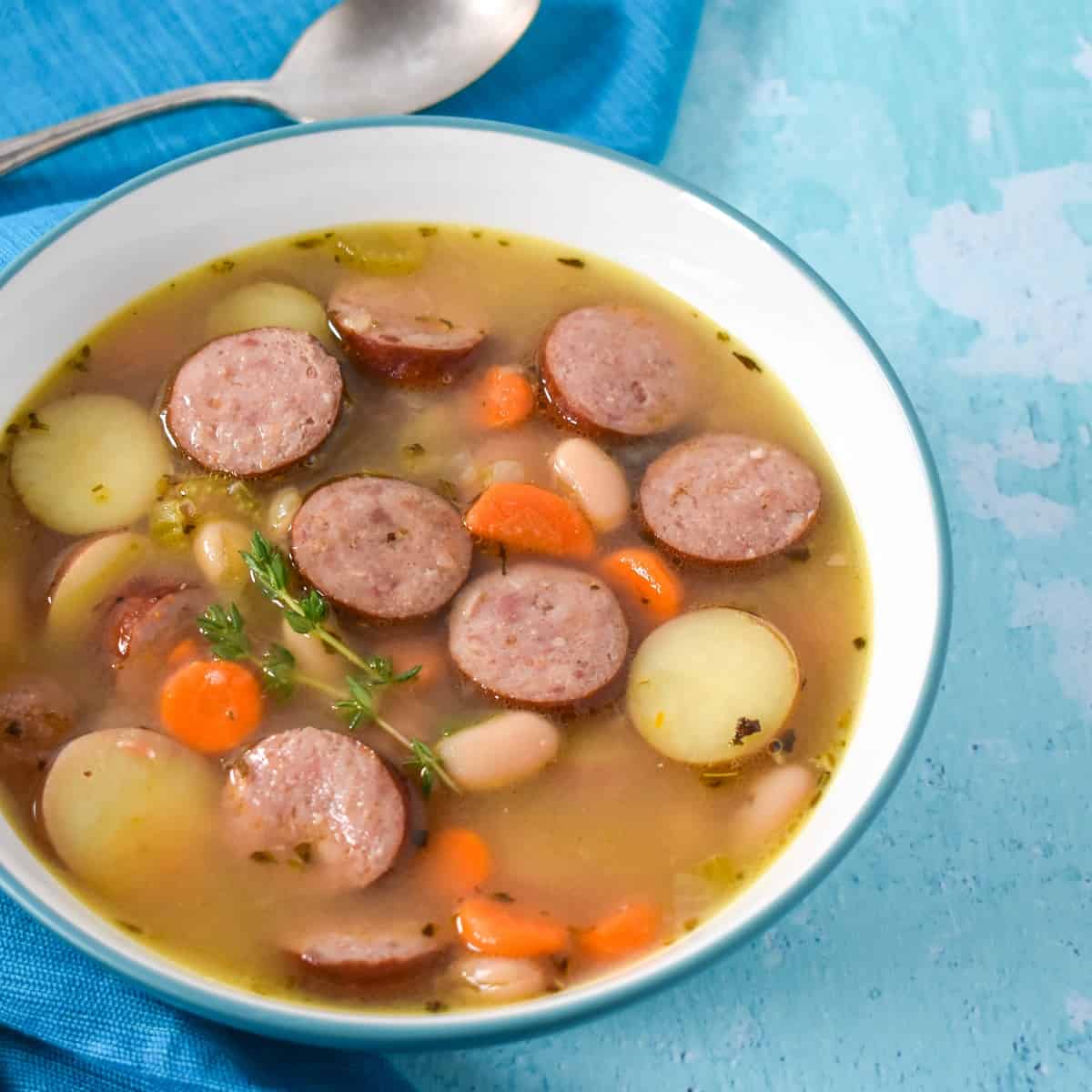 An image of the smoked sausage soup served in a white bowl with an aqua rim. The bowl is set on a light blue table with an aqua linen and spoon to the left side.