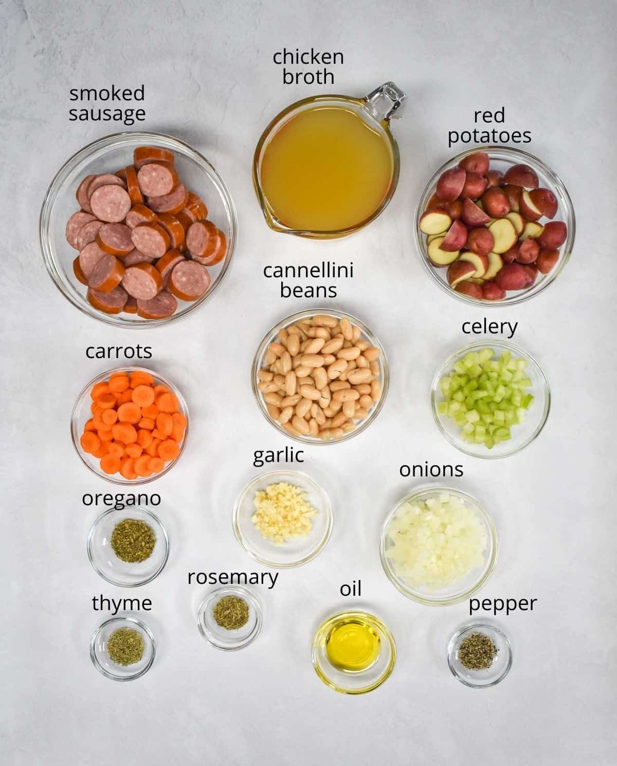 The ingredients for the sausage soup prepped and arranged in glass bowls on a white table. Each ingredient has a label with the name above it in small black letters.