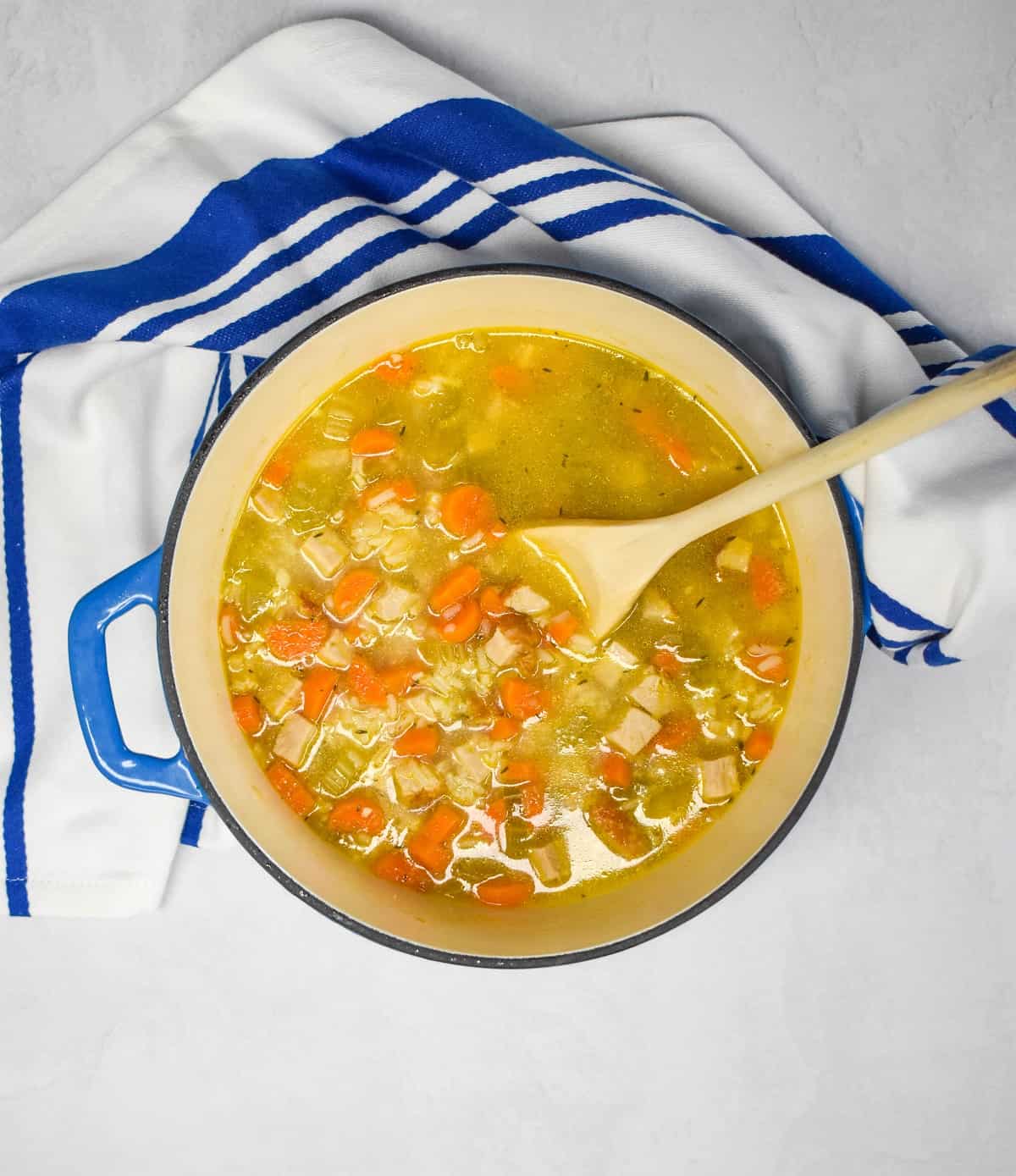 An image of the turkey and rice soup in a blue pot that is set on a white table with a blue and white kitchen towel and a wooden spoon in the pot.