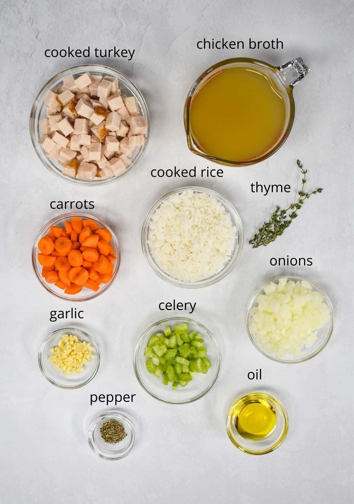 The ingredients for the soup prepped and arranged in glass bowls on a white table. Each ingredient has a label with the name above it in small black letters.