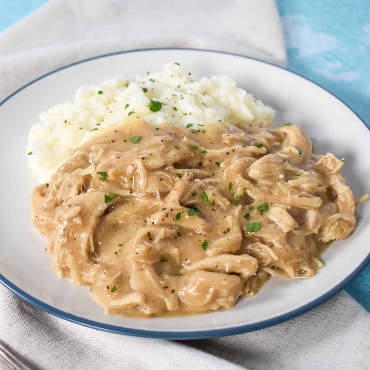 The chicken and gravy served with mashed potatoes on a white plate with a blue rim. The plate is set on a beige napkin on a light blue table.