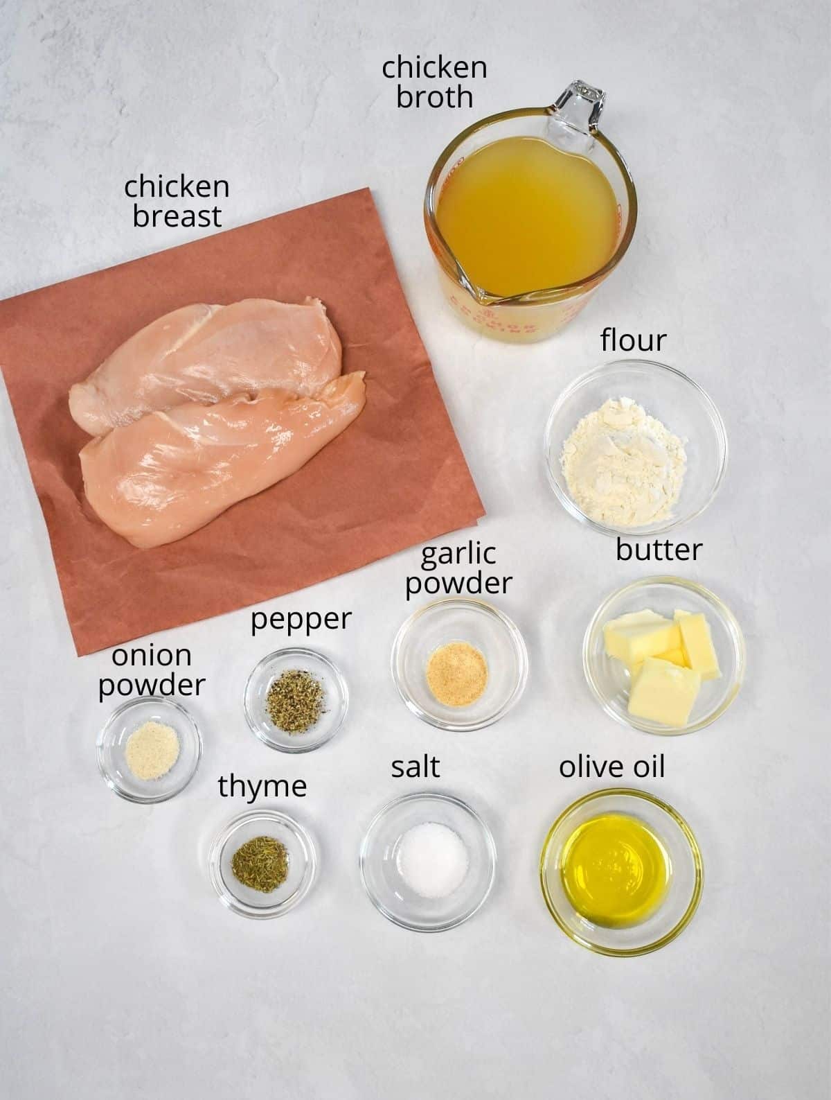 The ingredients for the dish prepped and arranged in glass bowls on a white table. The chicken is on butcher paper. Each ingredient has a label with the name above it in small black letters.