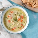 An image of the chicken pot pie soup served in a white bowl with an aqua rim. The bowl is set on an aqua colored table with an off-white linen to the left side an pie crust crackers on a wood cutting board to the top right side.