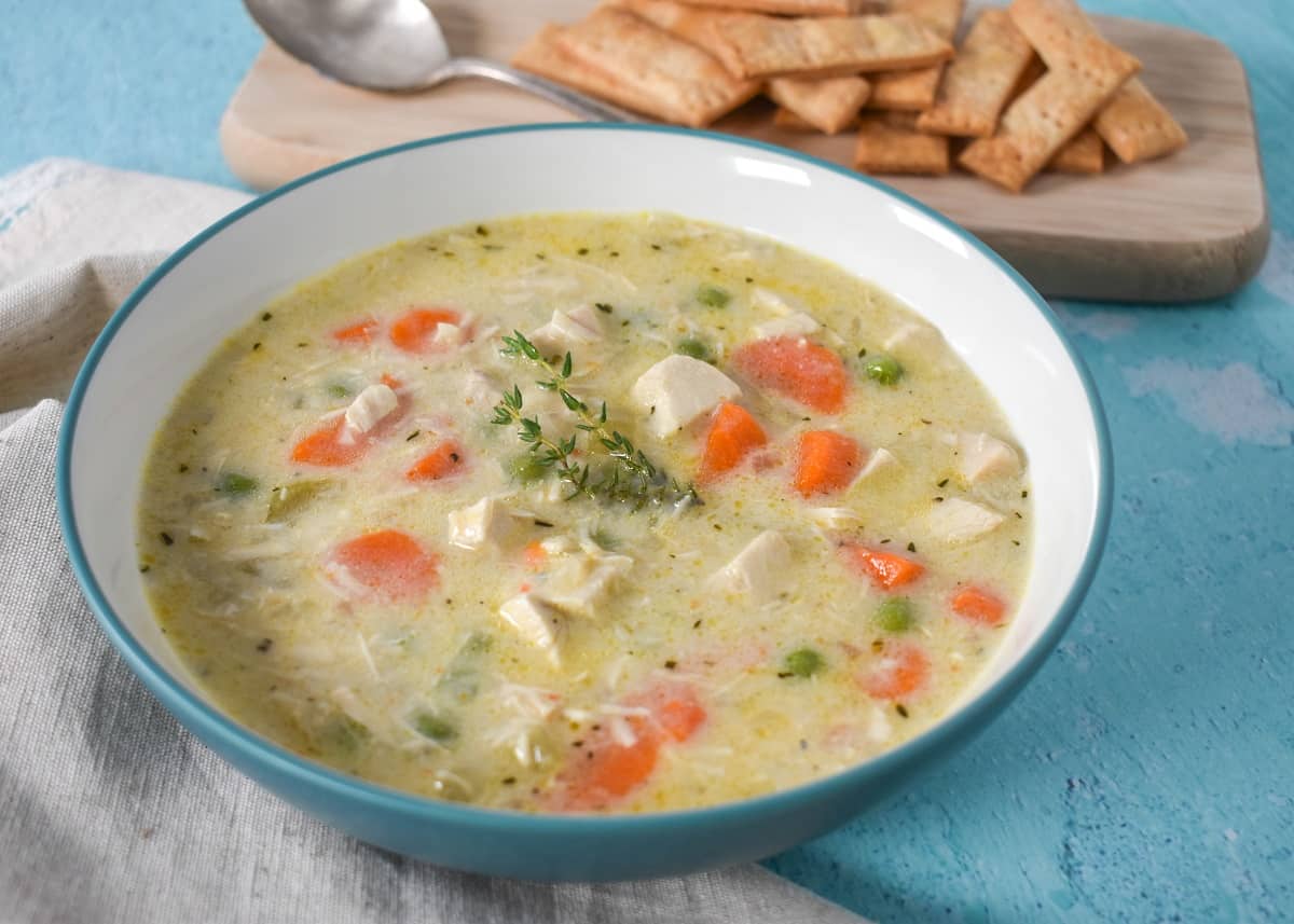 An image of the pot pie soup served in a white bowl with an aqua rim. The bowl is set on an aqua colored table with an off-white linen to the left side an pie crust crackers and a spoon on a wood cutting board to the top right side.