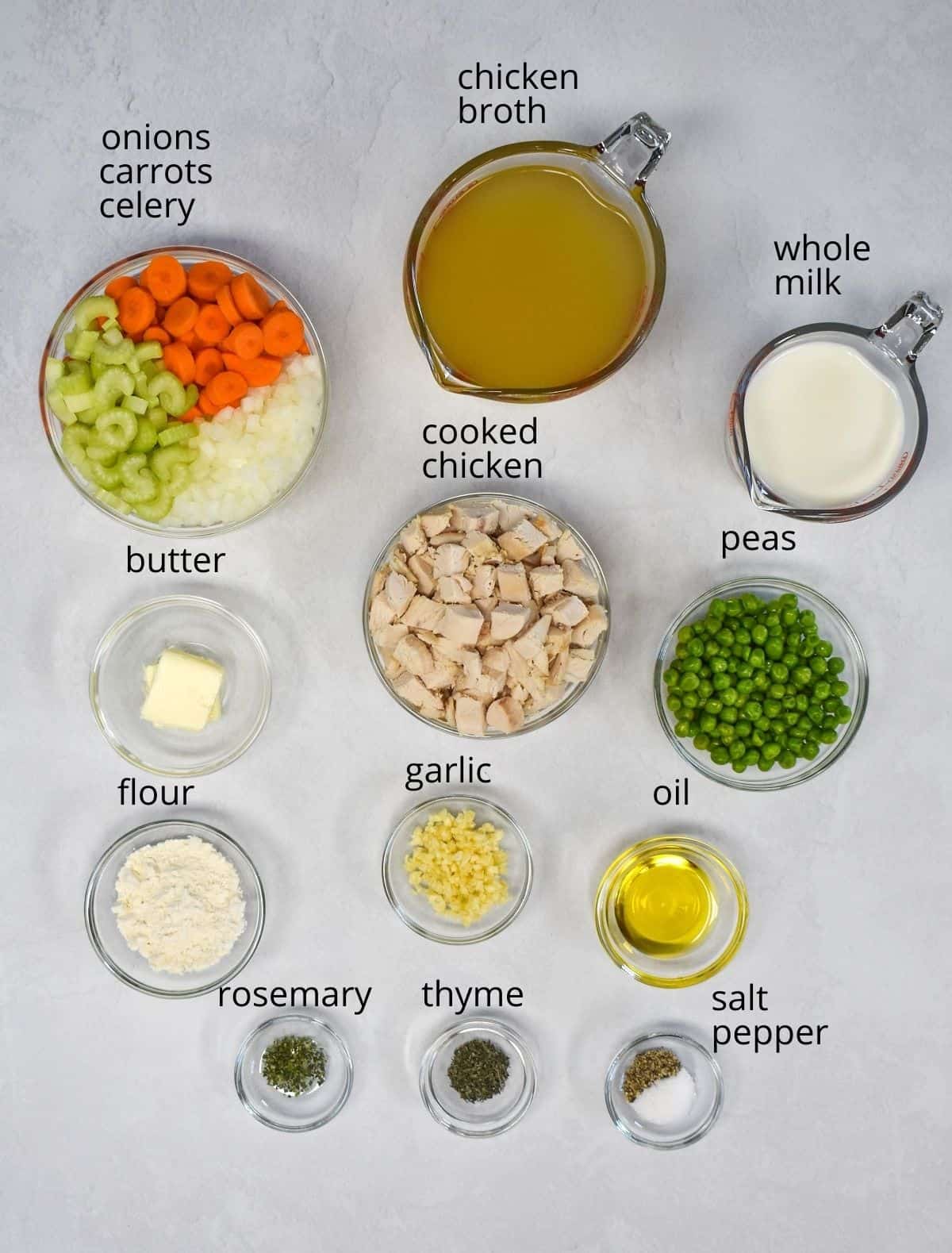The ingredients for the pot pie soup prepped and arranged in glass bowls on a white table. Each ingredient has a label with the name above it in small black letters.