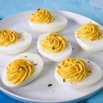 An image of six prepared deviled eggs garnished with a sprinkle of paprika and chopped parsley. The eggs are set on a white plated on an aqua table.