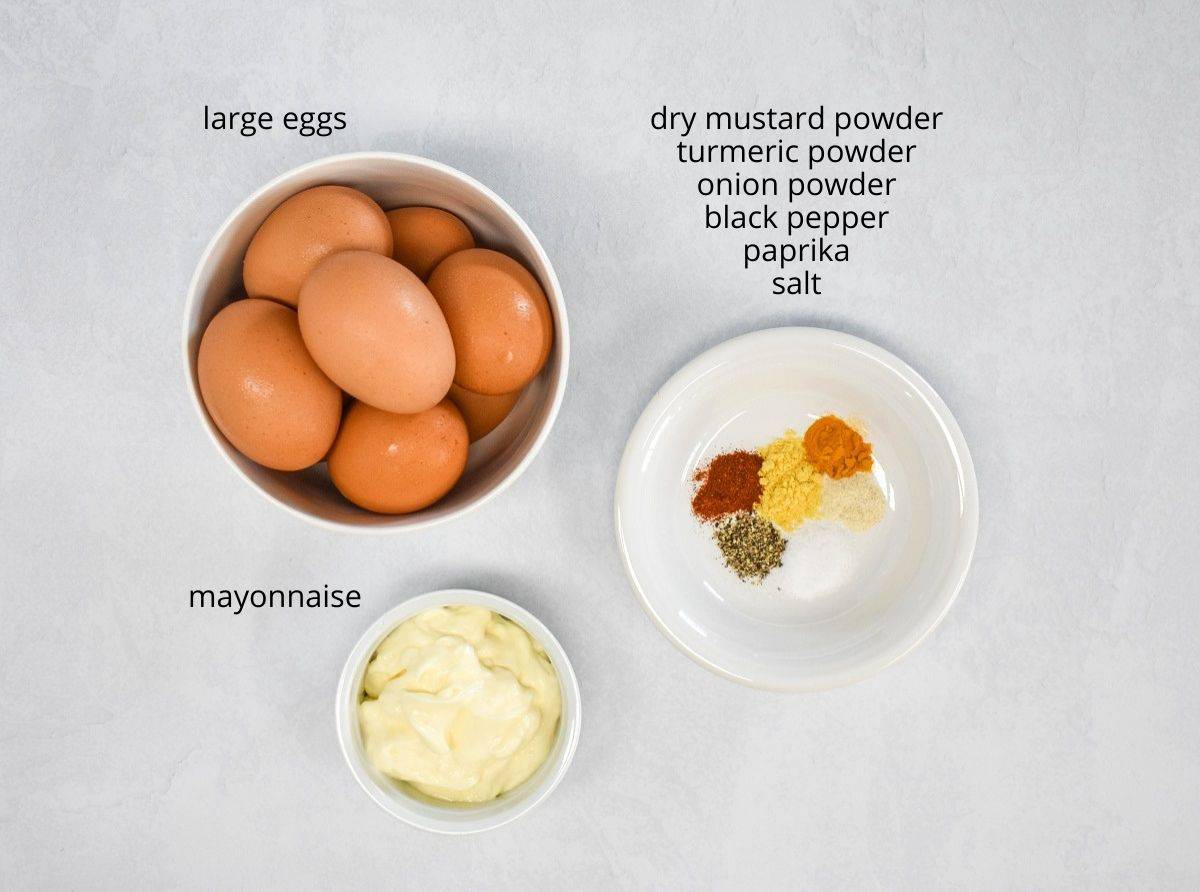 The ingredients for the deviled eggs arranged on a white table. The spices are all arranged on a white plate. They are labeled with small black letters.