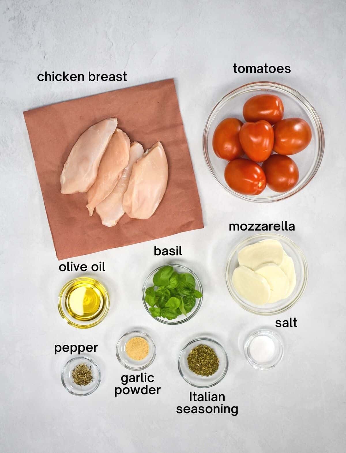 The ingredients for the dish arranged in glass bowls and set on a white table. The chicken is set on butcher paper. Each ingredient is labeled in small black letters.
