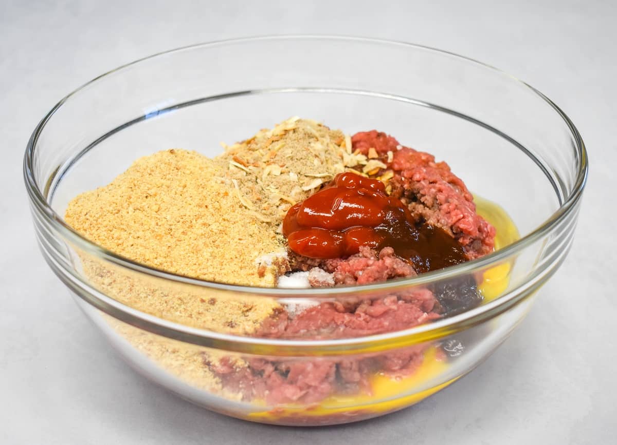 The ingredients for the mini meatloaves before mixing in a large, glass bowl that is set on a white table.