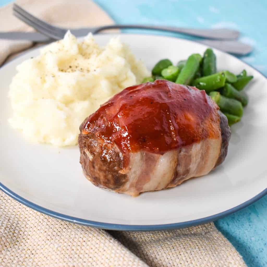 An image of the finished mini meatloaf served with mashed potatoes and green beans on a white plate with a blue rim. The plate is set on a light blue table with a beige linen and silverware in the background.