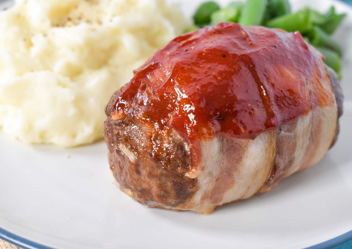 A close up image of a mini meatloaf served with mashed potatoes and green beans, set on a white plate with a blue rim.