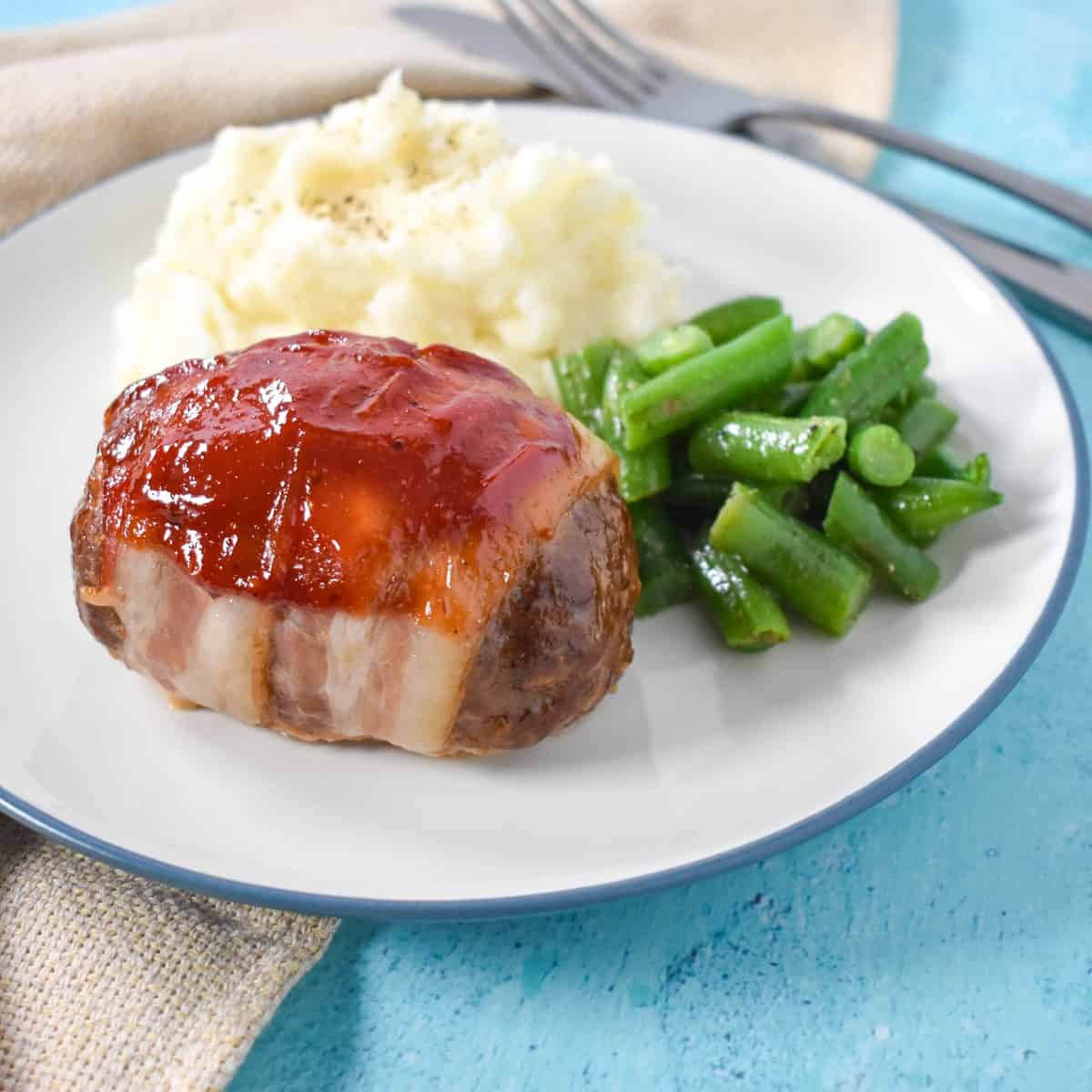 An image of the finished meatloaf served with mashed potatoes and green beans on a white plate with a blue rim. The plate is set on a light blue table with a beige linen and silverware in the background.