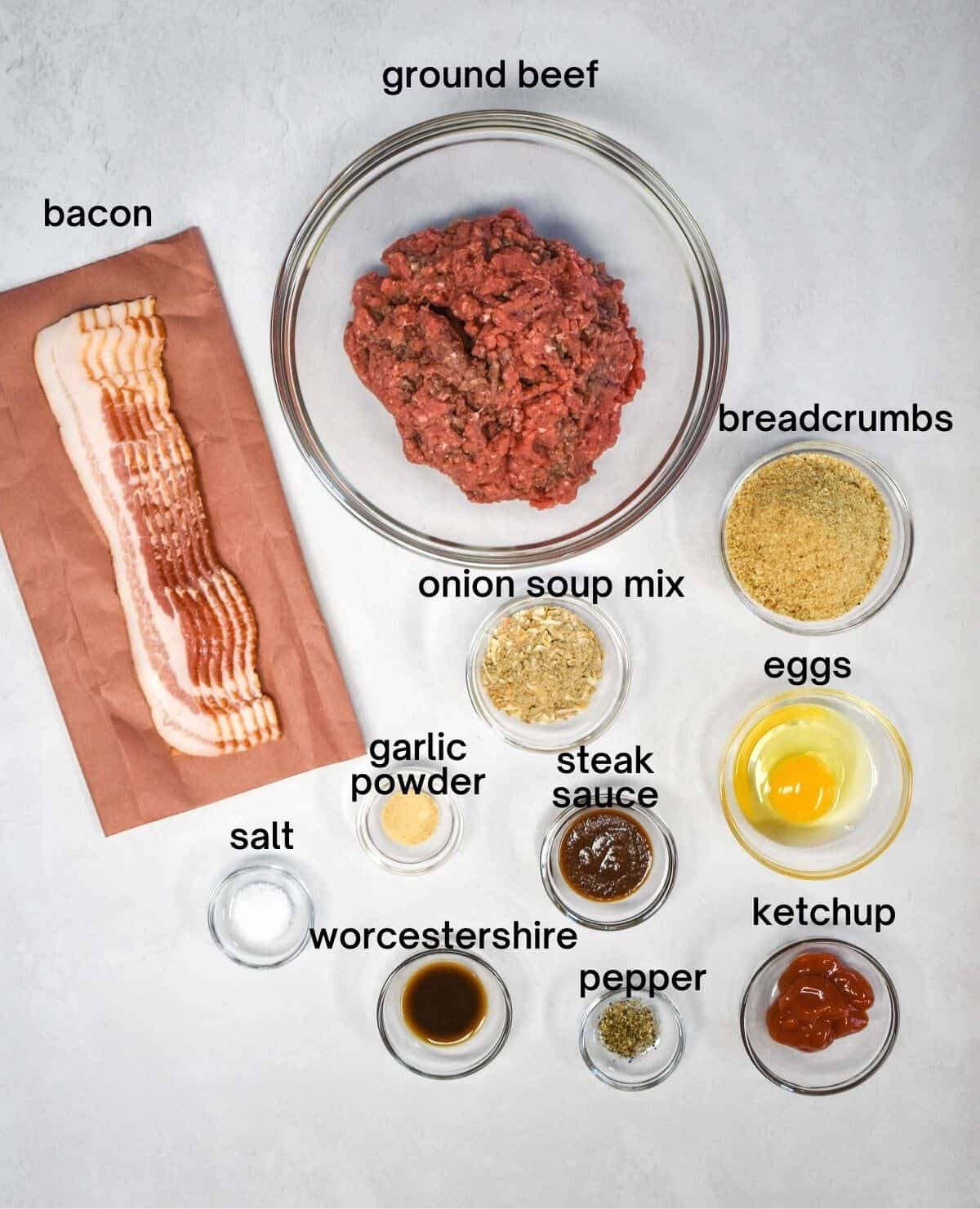The ingredients for the meatloaf arranged in glass bowls on a white table. Each ingredients has the name above it in black letters.
