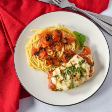 The baked chicken caprese served with a side of pasta topped with tomatoes, served on a white plate with a black rim. The plate is set on a white table with a red linen.