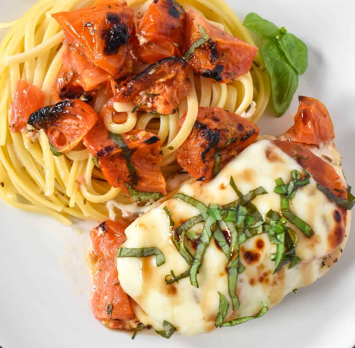 A close up of the caprese chicken served with pasta that is topped with the tomatoes on a white plate.