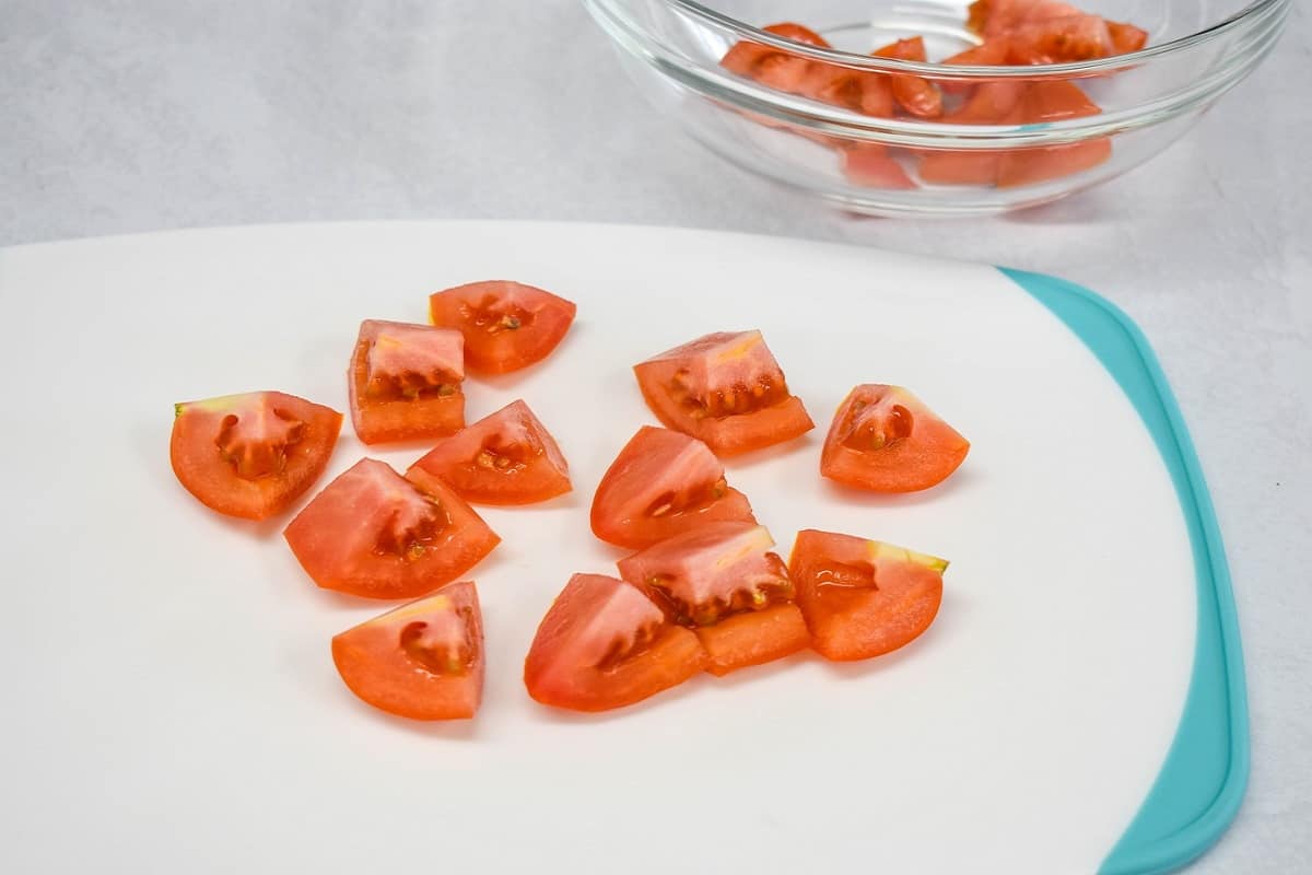 Cut tomatoes on a white and teal cutting board with a clear bowl in the background with some cut tomatoes in it.