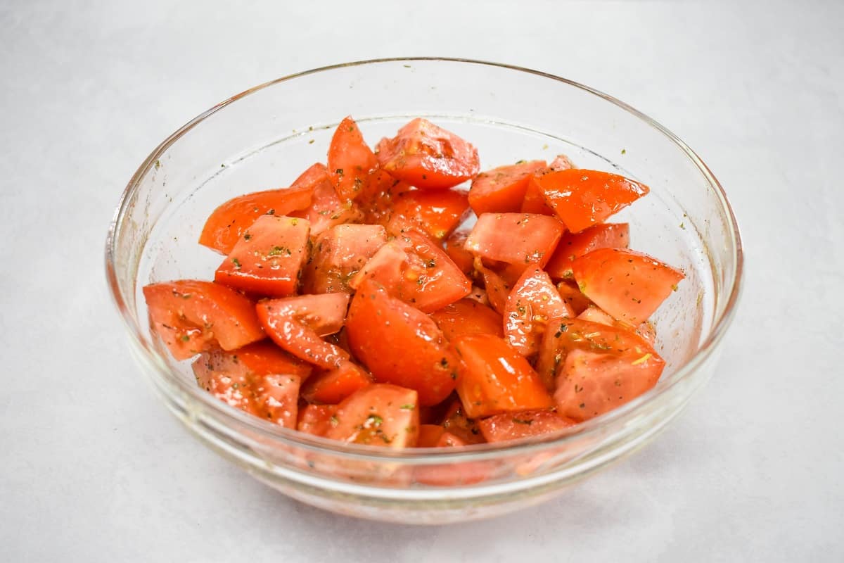 Seasoned cut tomatoes in a large, clear bowl set on a white table.