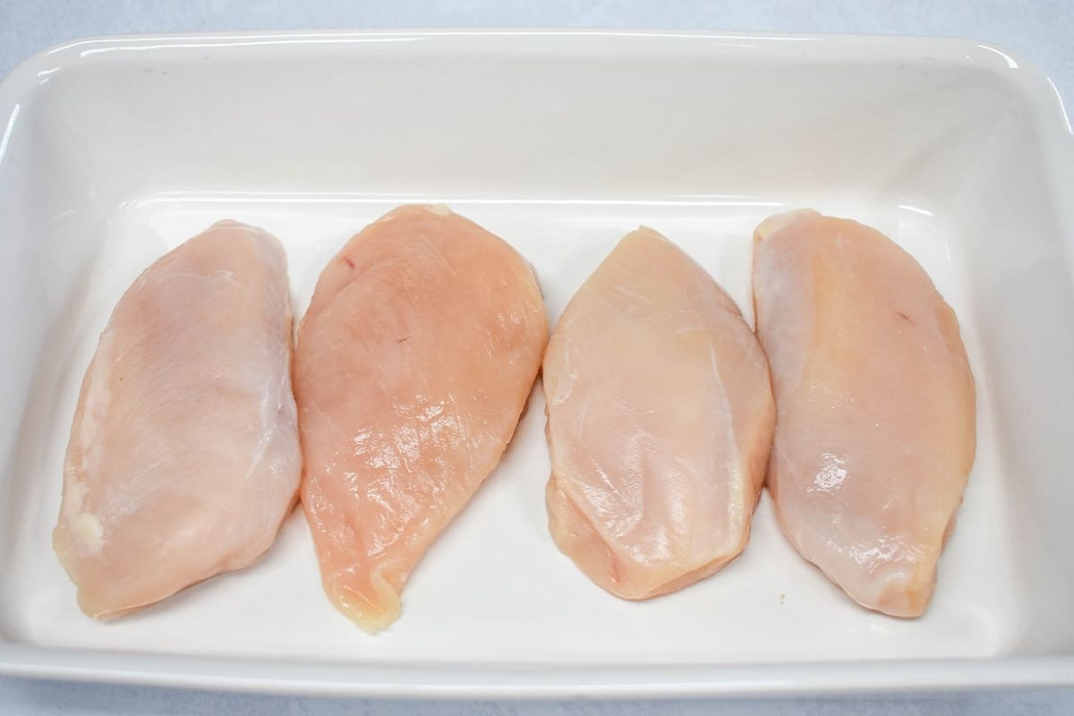 Four pieces of chicken breast in a white baking dish.