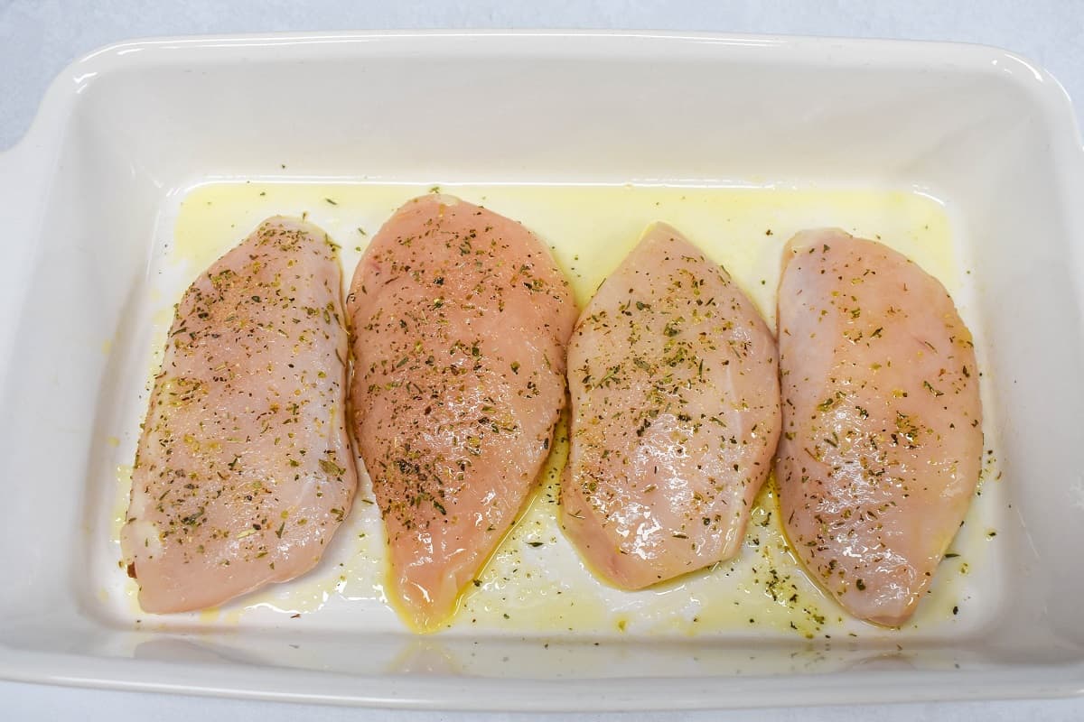 Four pieces of chicken breast covered with olive oil and seasoned in a white baking dish.