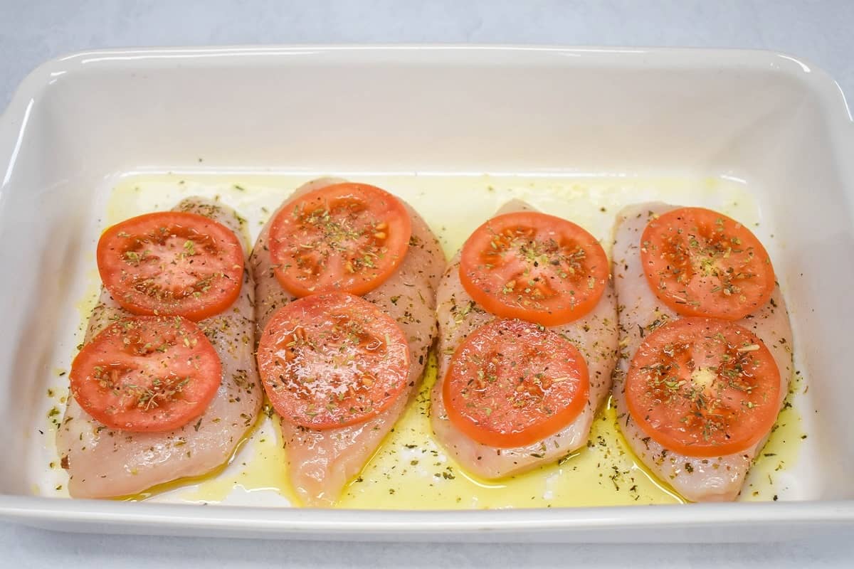 Four seasoned chicken breast pieces topped with tomatoes slices and olive oil in a white baking dish.